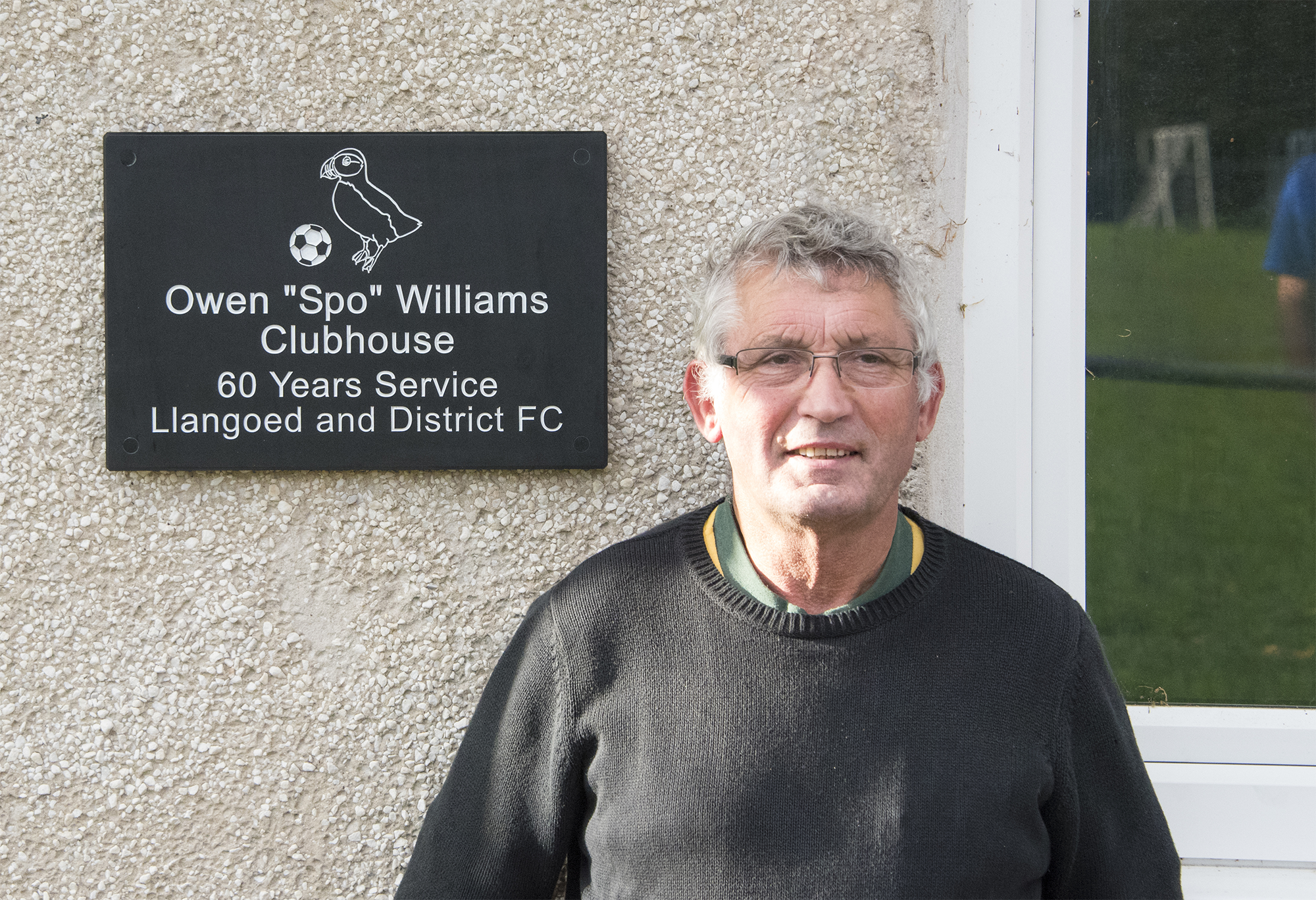 Owen Spo Williams standing beside the slate plaque naming the Owen Spo Williams Clubhouse, marking 60 years of service to Llangoed and District FC